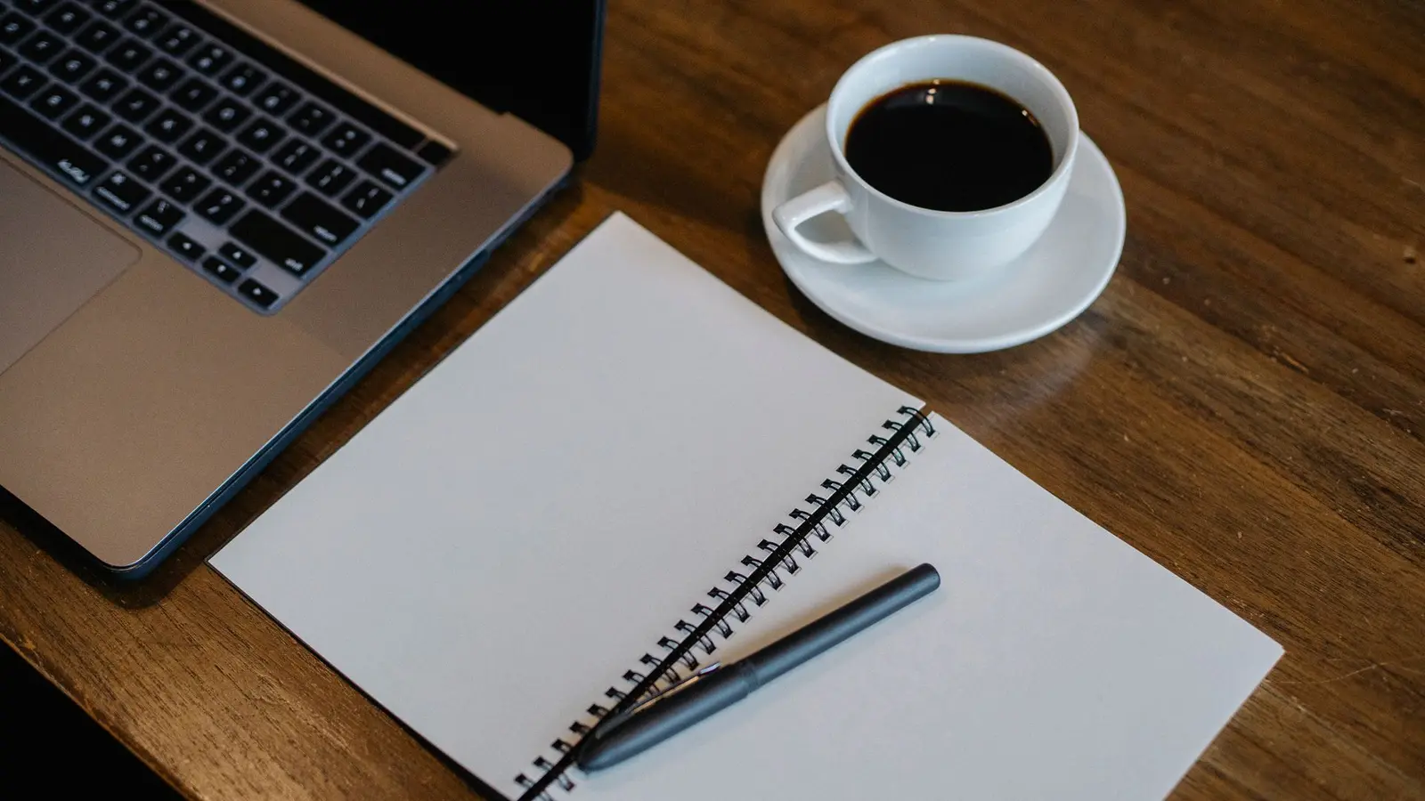 Buku catatan, pena, dan secangkir kopi di samping laptop